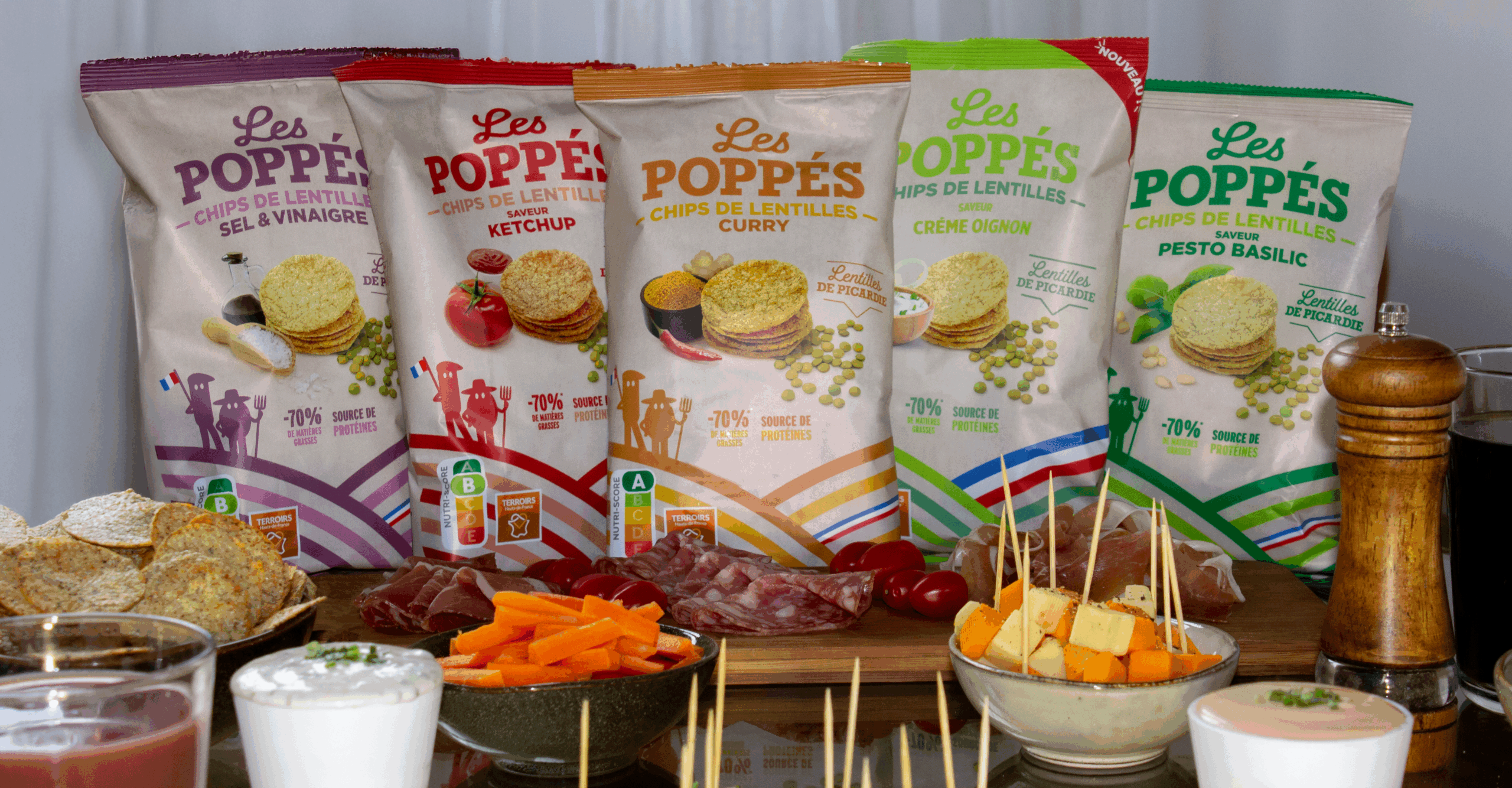Cinq paquets de chips de lentilles Les Poppés (Sel & Vinaigre, Ketchup, Curry, Crème Oignon, Pesto Basilic) posés sur une table garnie de fromage, charcuterie, légumes et sauces pour un apéritif gourmand et convivial.