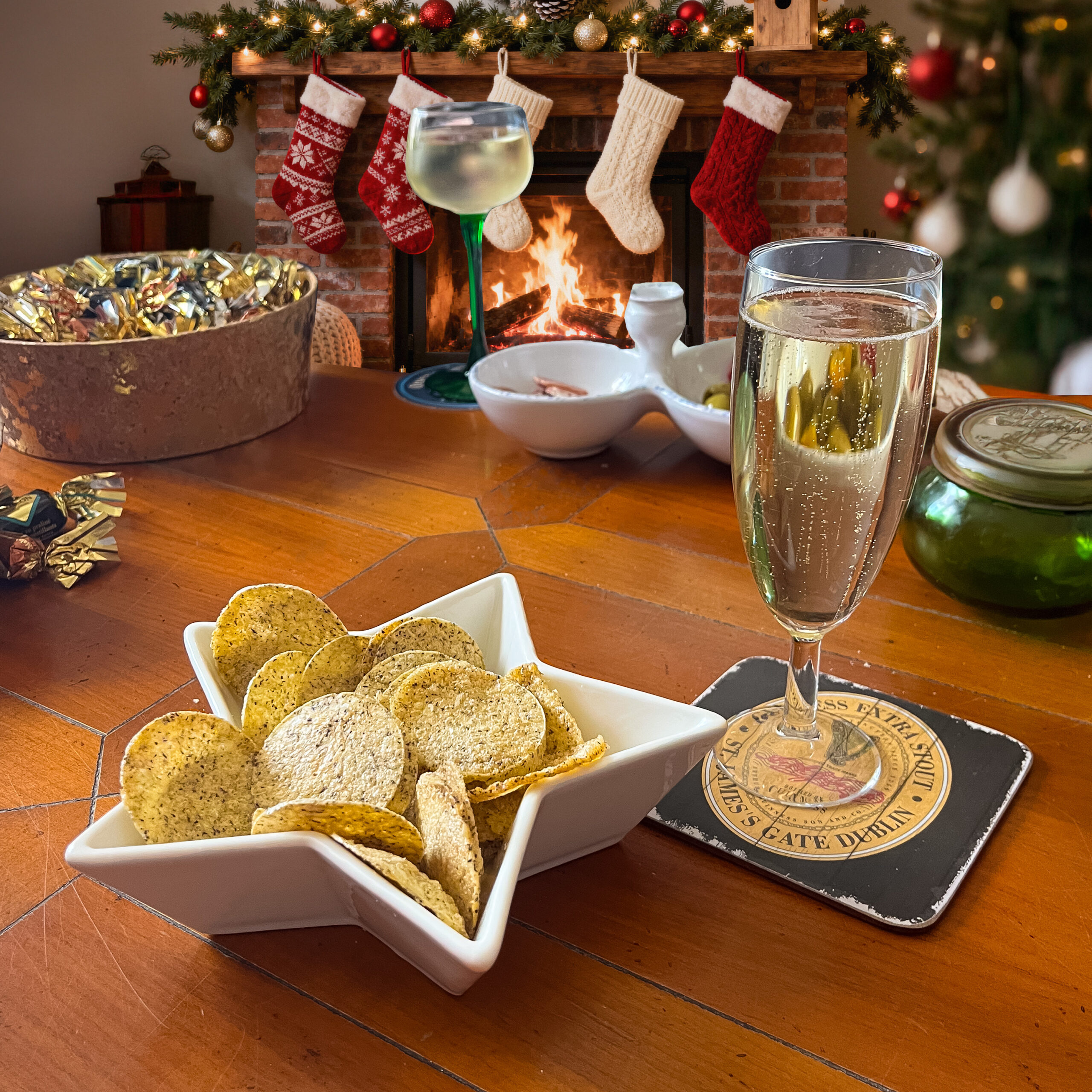 photo ambiance de noel : des chips disposées dans un bol blanc en forme d'étoile avec une coupe de champagne à droite et des papillotes à gauche et une cheminée avec des chaussettes de noel accrochées sur la devanture en arrière plan